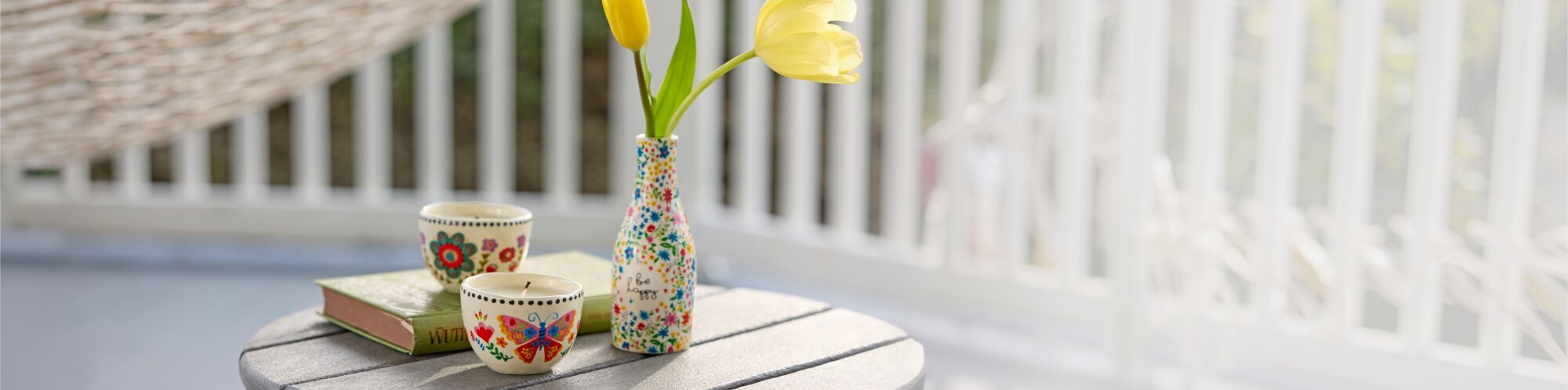 Sunlit porch scene with a small round table holding a Natural Life floral vase with yellow tulips, two patterned ceramic cups (one with a butterfly design), and a stacked book, set against white railing in soft focus.