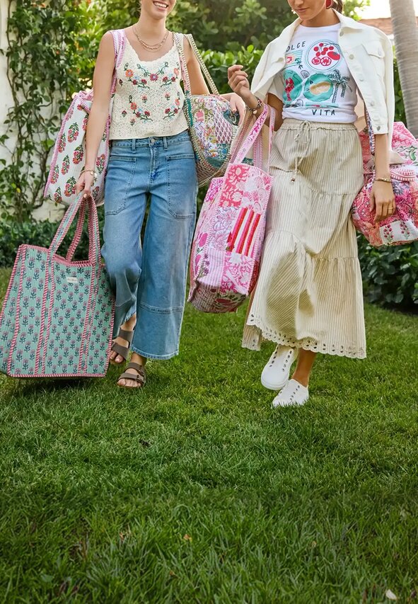 Two women walk through a lush garden courtyard carrying colorful Strawflower tote bags and duffels. One wears a floral crochet tank with wide-leg denim jeans, while the other wears a graphic “Dolce Vita” tee with a striped midi skirt and white jacket draped over her shoulders. The scene is bright and sunlit with greenery, palm trees, and a Mediterranean-inspired home in the background.