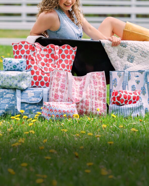 Woman sitting on grass behind a collection of floral and patterned Strawflower tote bags, duffels and cosmetic bags.