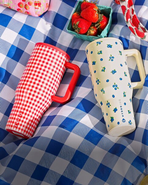 Patriotic Hydrojug drinkware and picnic accessories on a blue gingham blanket, including tumblers, a red gingham cooler, and strawberries.