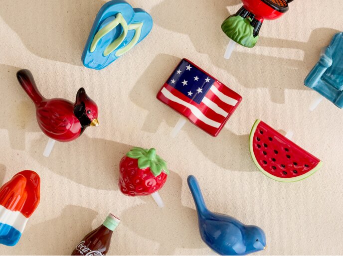 Image of various Nora Fleming mini ornaments, featuring blue flip flops, an American flag, a watermelon slice, a cardinal, a strawberry, a blue bird, a red white and blue popsicle, and a bottle of Coca Cola.