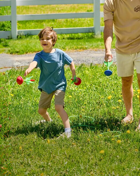Child and adult playing an outdoor lawn game in a sunny grassy field beside a white fence.