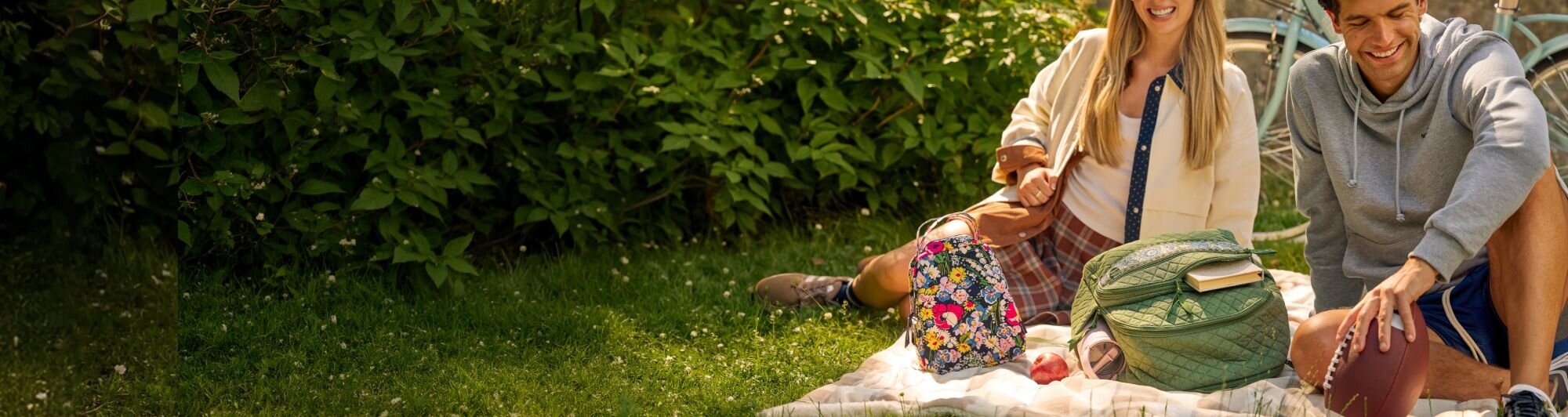 Person lying on green grass dotted with yellow flowers, wearing a white T-shirt, light-wash jeans with embroidered daisies, and clear shoes. A large Vera Bradley duffel bag in Poppy Fields and matching keychain pouch rest beside them, creating a colorful spring-inspired outdoor scene.