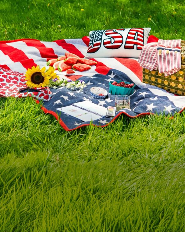 Patriotic picnic setup with an American flag blanket, strawberries, watermelon, flowers, and woven picnic basket spread across green grass.