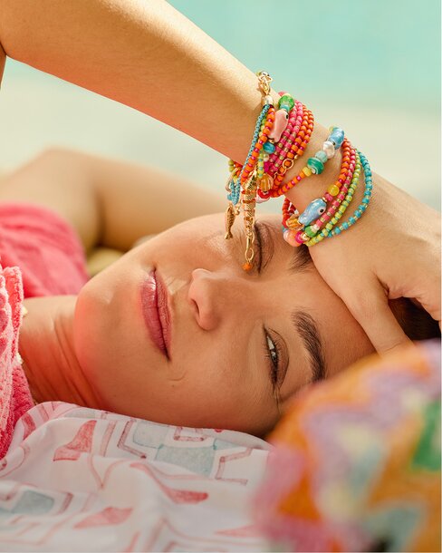 A close-up portrait shows a woman lounging poolside with her arm resting across her forehead, wearing stacked colorful beaded bracelets with charms in bright pink, orange, turquoise, and gold tones. Sunlight highlights her relaxed expression and soft makeup, creating a vibrant, carefree summer mood.
