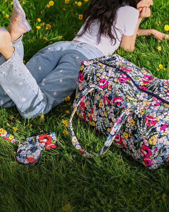 A woman lounges in the grass wearing embroidered light-wash jeans beside a colorful floral Vera Bradley duffel bag and matching pouch.