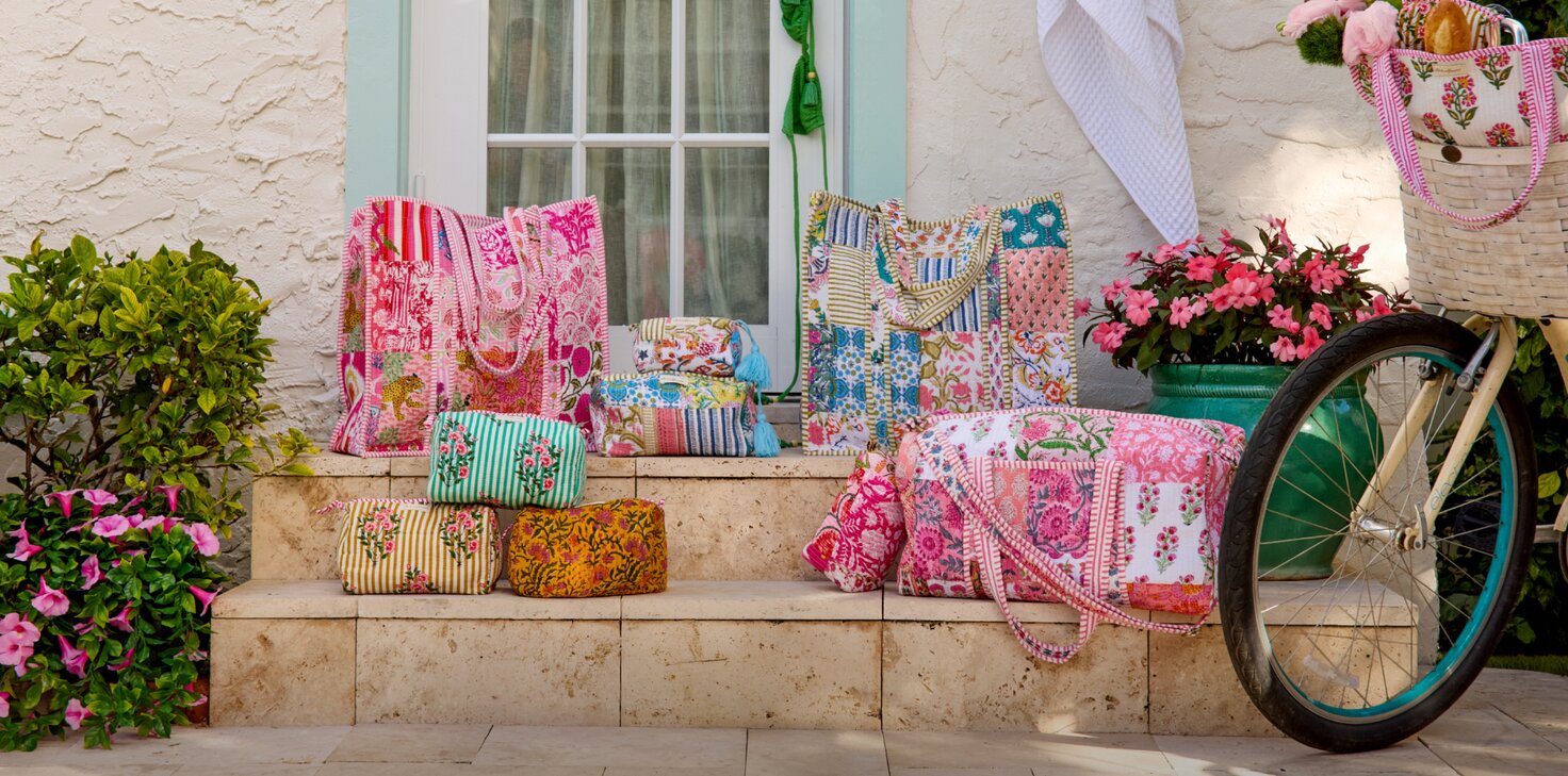 Colorful Strawflower tote bags, duffels and matching pouches arranged on stone steps outside a pastel home, surrounded by blooming flowers, with a bicycle and basket filled with pink blooms nearby.