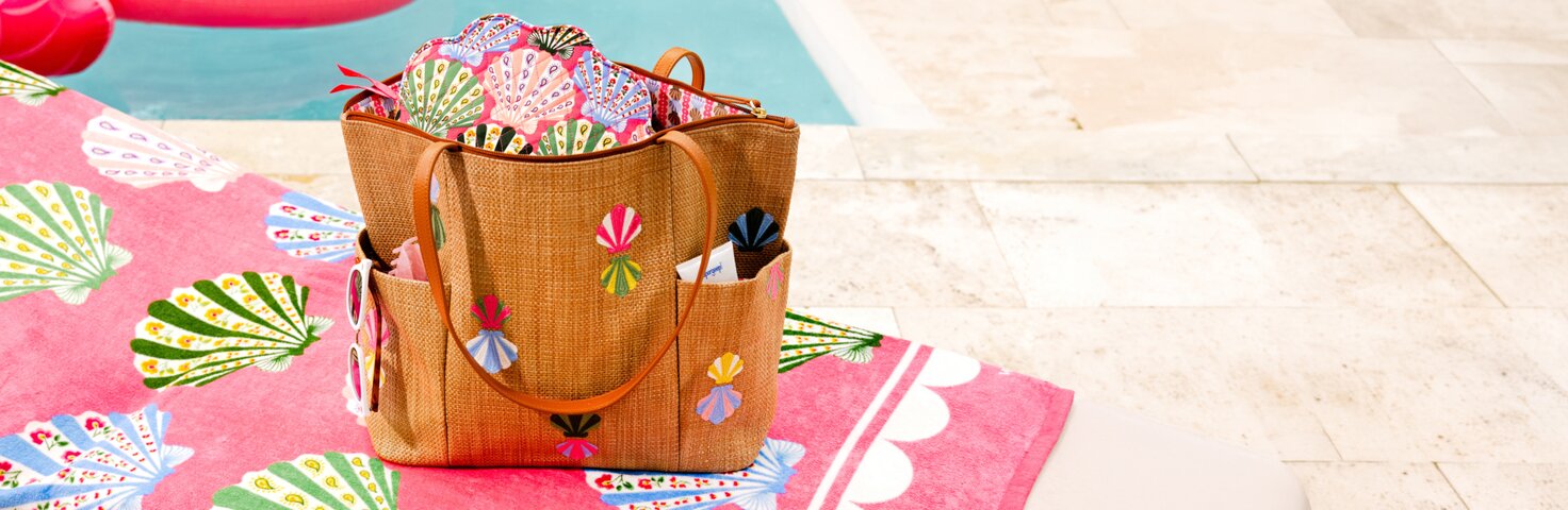 Vera Bradley faux straw beach tote with colorful embroidered shell details sitting on a Vera Bradley pink shell patterned beach towel beside a pool. The open tote is filled with matching printed accessories, with sunglasses tucked into the side pocket and a bright, resort-style poolside setting in the background.