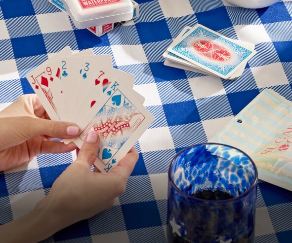 Hand holding playing cards over a blue gingham tablecloth with card games and picnic accessories.