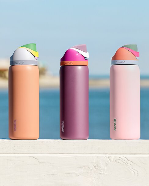 Three pastel-colored Owala insulated water bottles lined up on a white ledge with a blurred beach and ocean in the background.