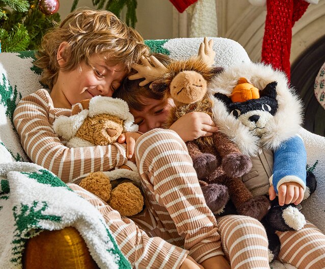 Two children in matching striped pajamas cuddle on a cozy couch with Jellycat plush toys, surrounded by festive holiday blankets.