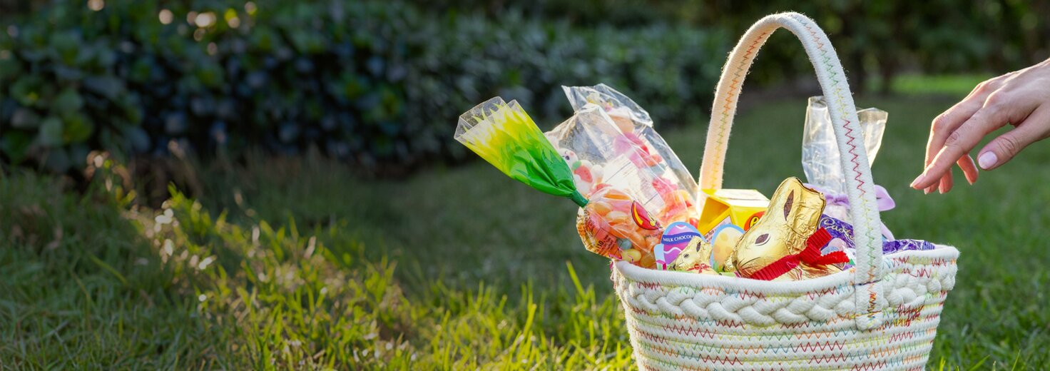 Woven Easter basket sitting on grass, filled with colorful candy, chocolate bunnies, and packaged treats, as a hand reaches in from the side.