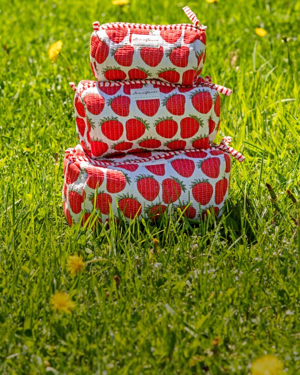 Strawberry-print quilted Strawflower set of 3 cosmetic pouches and fresh strawberries arranged in tall green grass on a bright summer day.