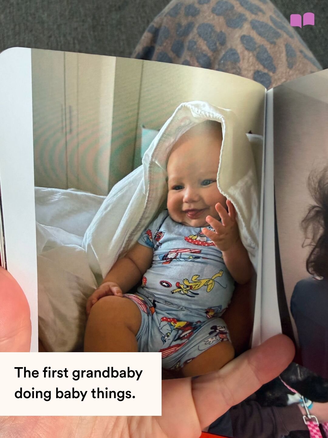 A Monthly Mini photo book page showing a smiling baby in a Disney onesie peeking from under a blanket, captioned about the first grandbaby