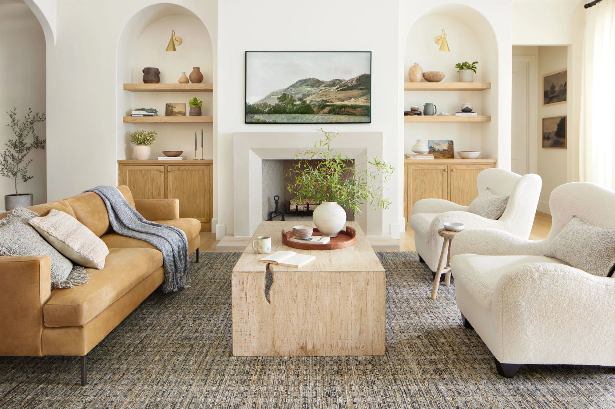 Inviting living room with a tan leather sofa, two white bouclé chairs and a wooden coffee table sitting on top of a rug in front of a fireplace.