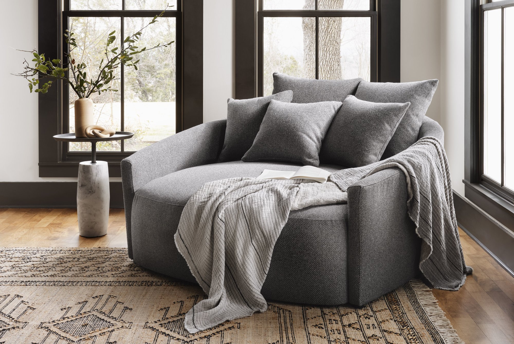 Spacious gray lounge chair styled with throw pillows and a cozy throw blanket sitting next to a side table in front of three large windows.