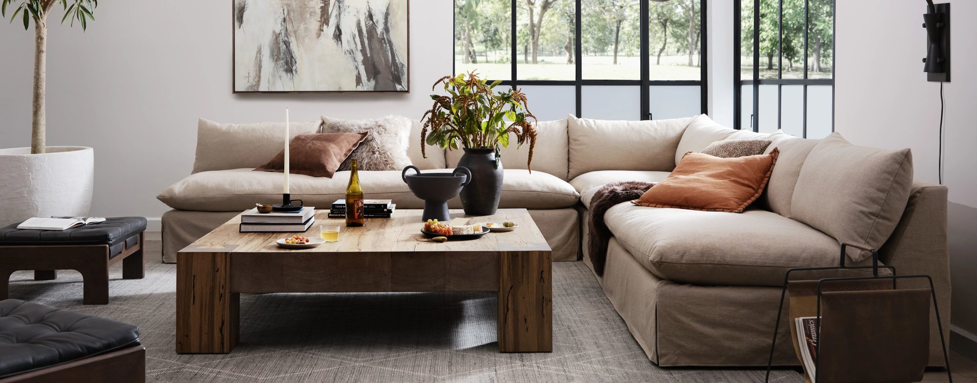 Wabi-sabi style living room with a linen sectional and wooden coffee table with natural wood variations sitting in front of large windows.