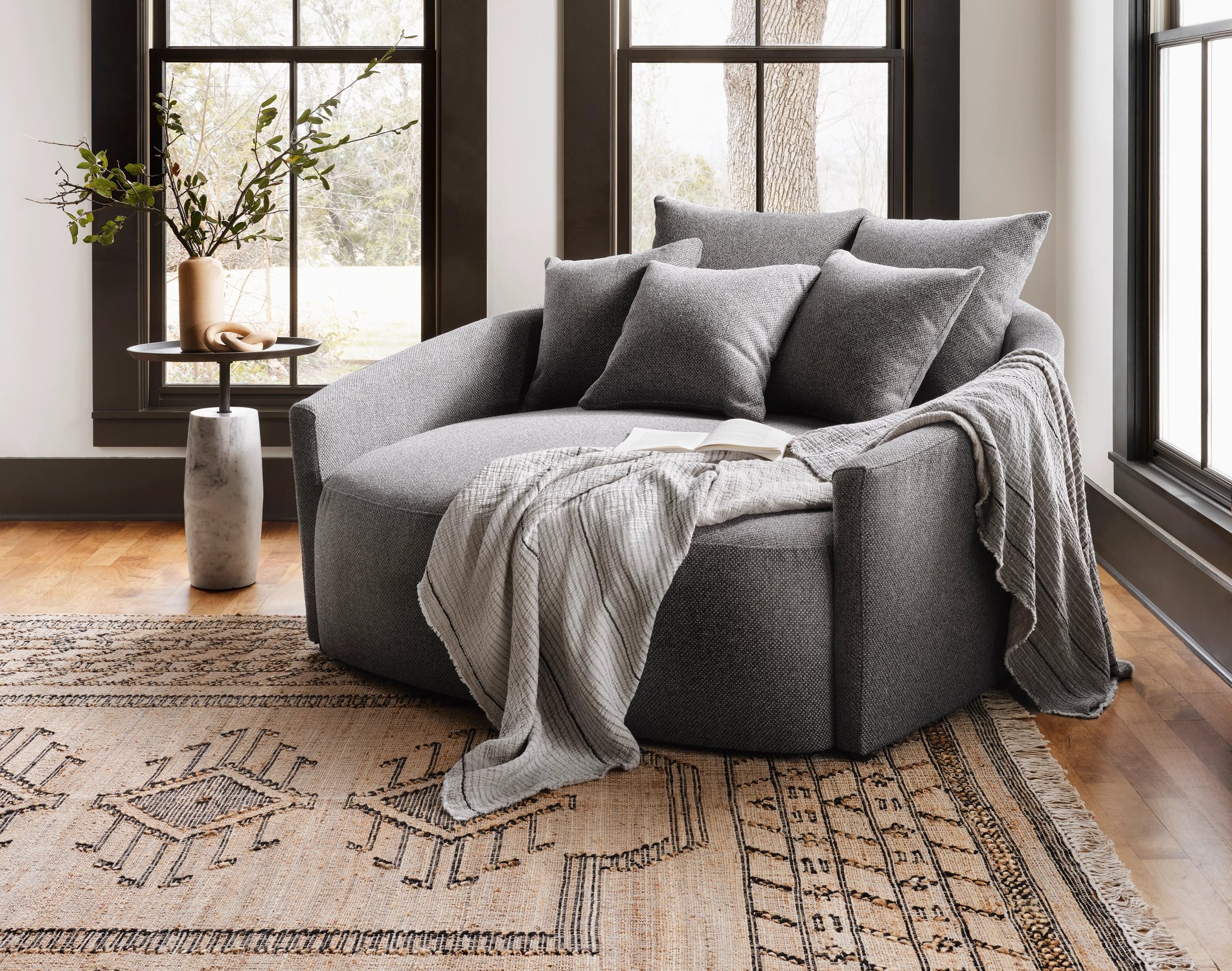 Spacious gray lounge chair styled with throw pillows and a cozy throw blanket sitting next to a side table in front of three large windows.