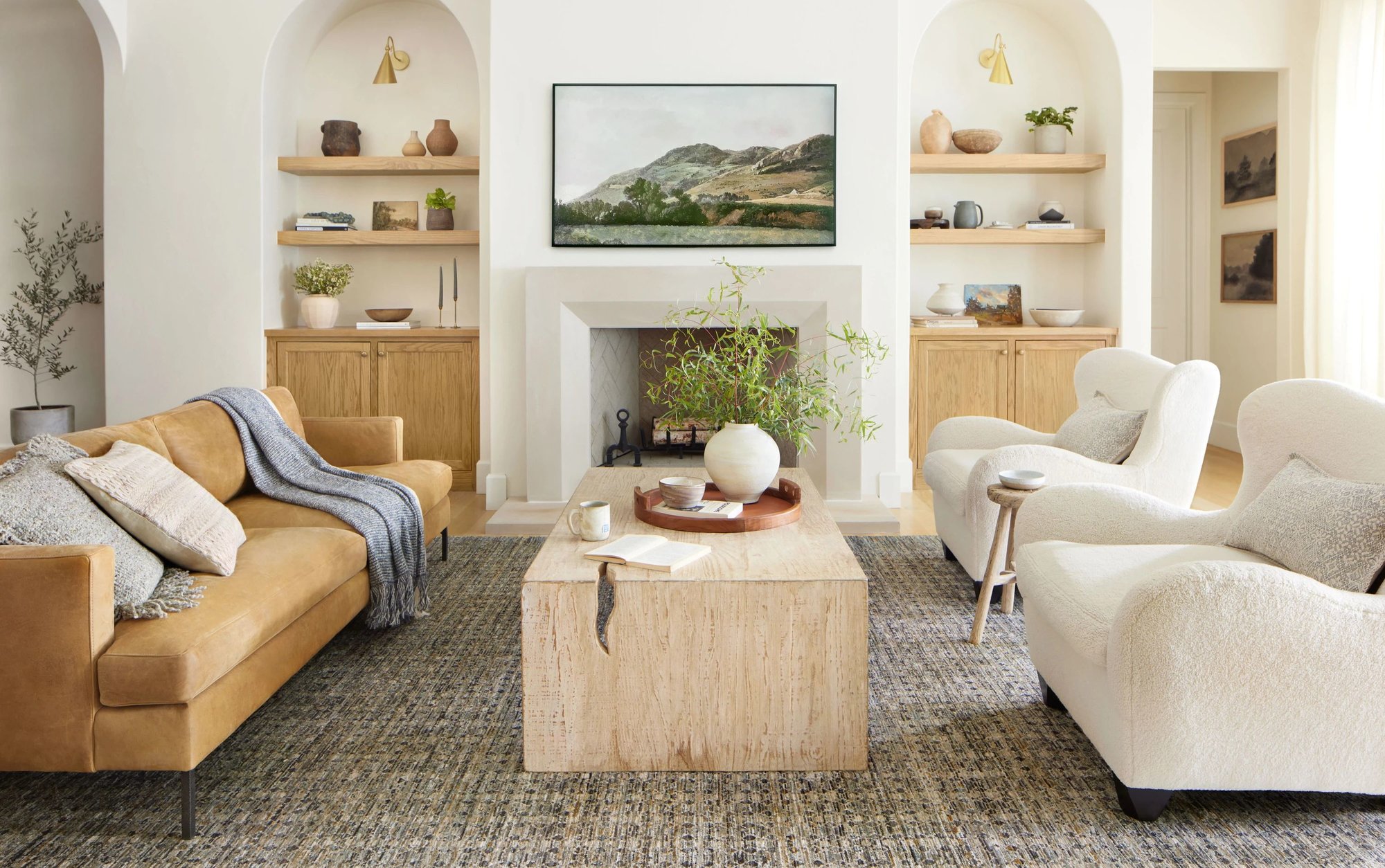 Inviting living room with a tan leather sofa, two white bouclé chairs and a wooden coffee table sitting on top of a rug in front of a fireplace.