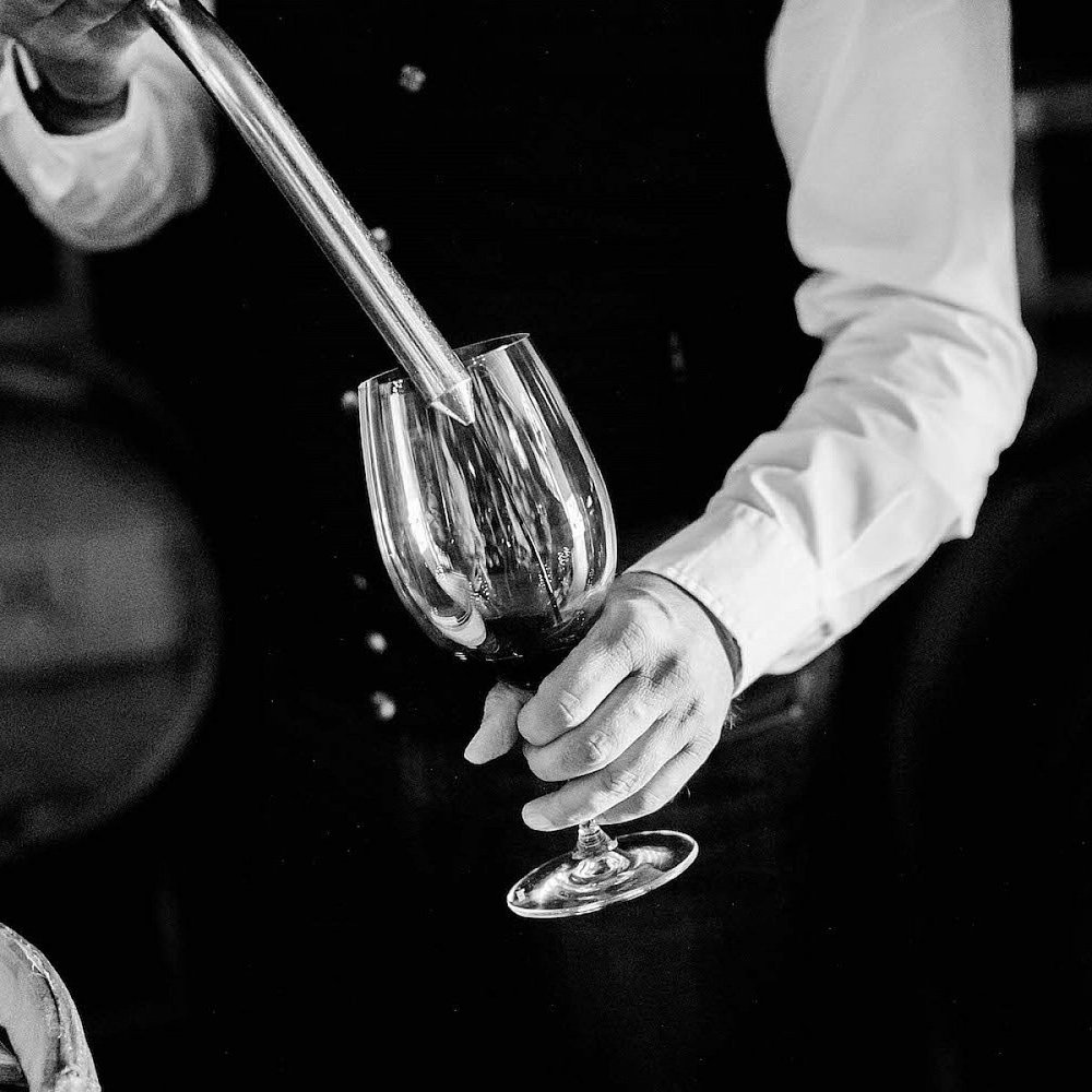 A winemaker tests wine from a barrel in a wine glass