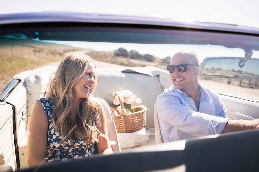 A couple drives in a convertible with a picnic basket in the backseat