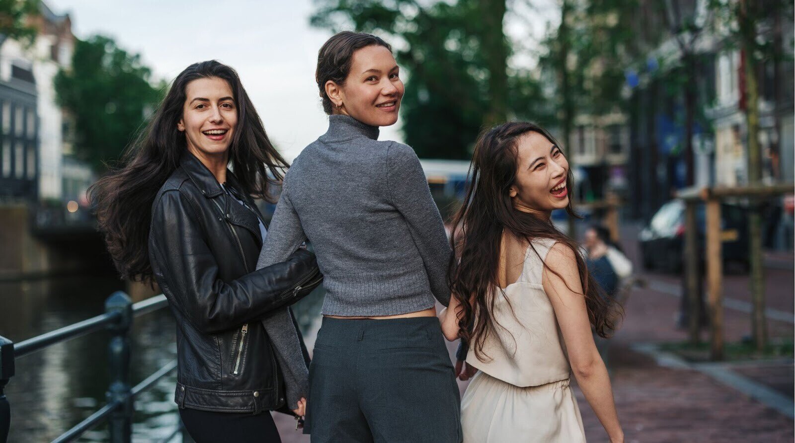 Três mulheres jovens andando pelas ruas de Amsterdã. Uma com jaqueta de couro e calça escura, outra com suéter e saia, e outra com macacão leve de mangas curtas.