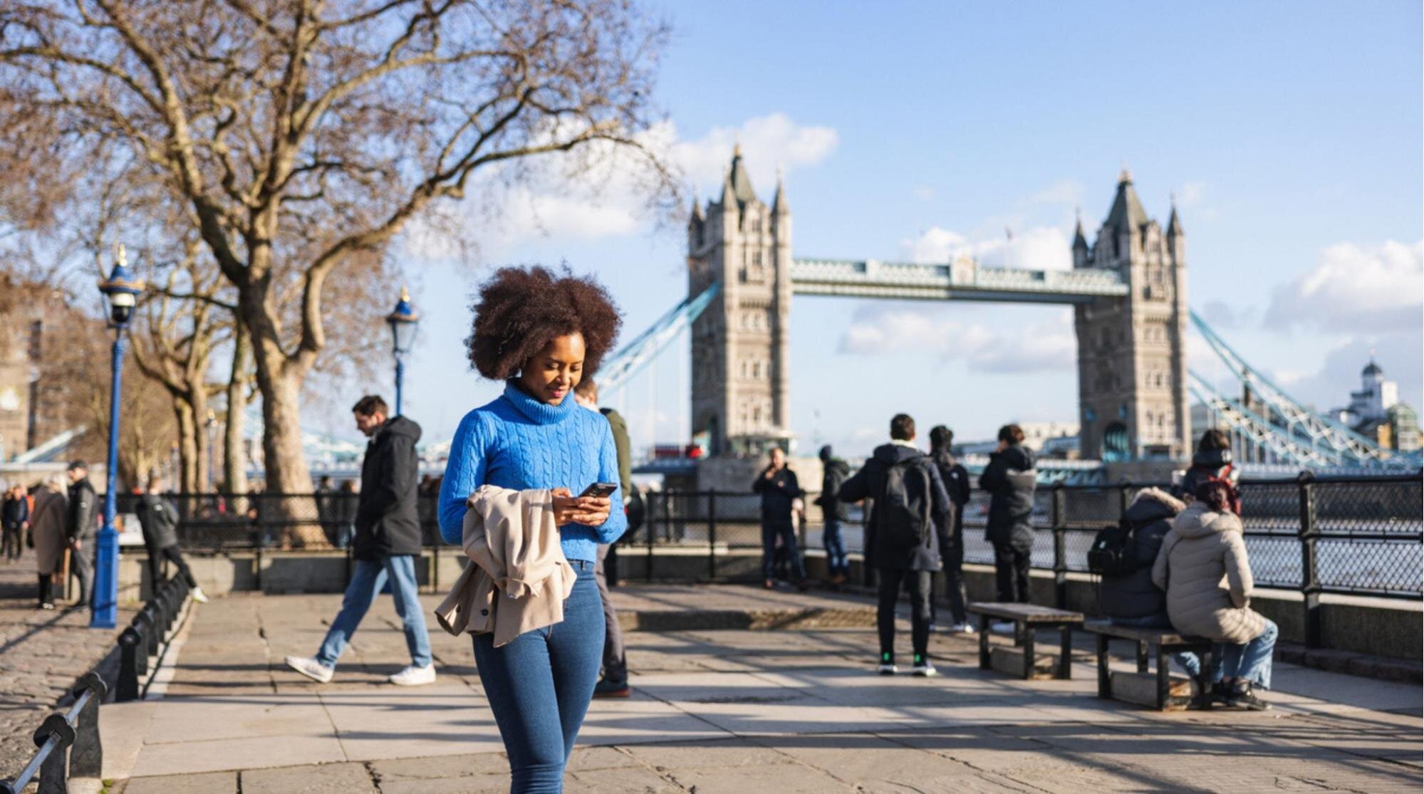 Turista caminhando às margens do Rio Tâmisa enquanto mexe no celular, com a Tower Bridge ao fundo. 