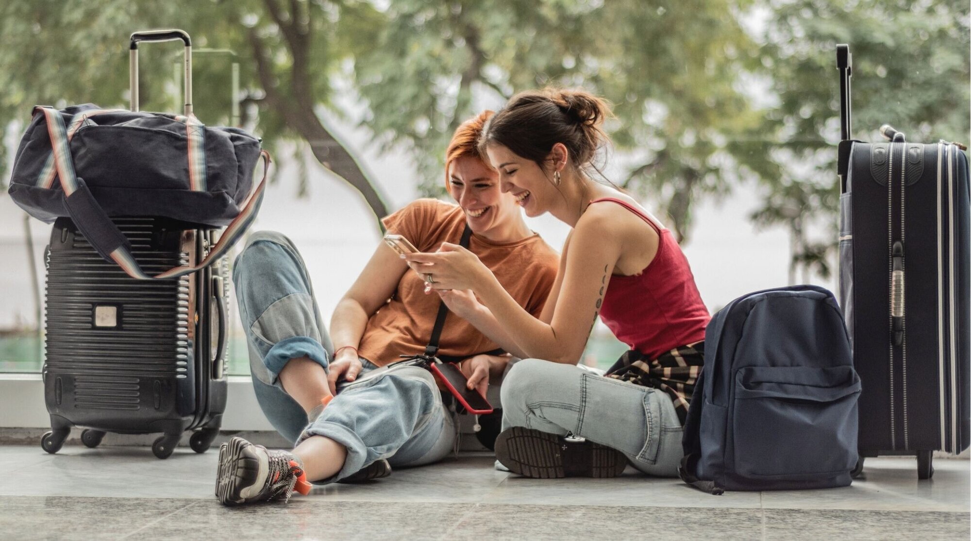 Jovens amigas usando o celular sentadas no chão enquanto esperam no aeroporto.
