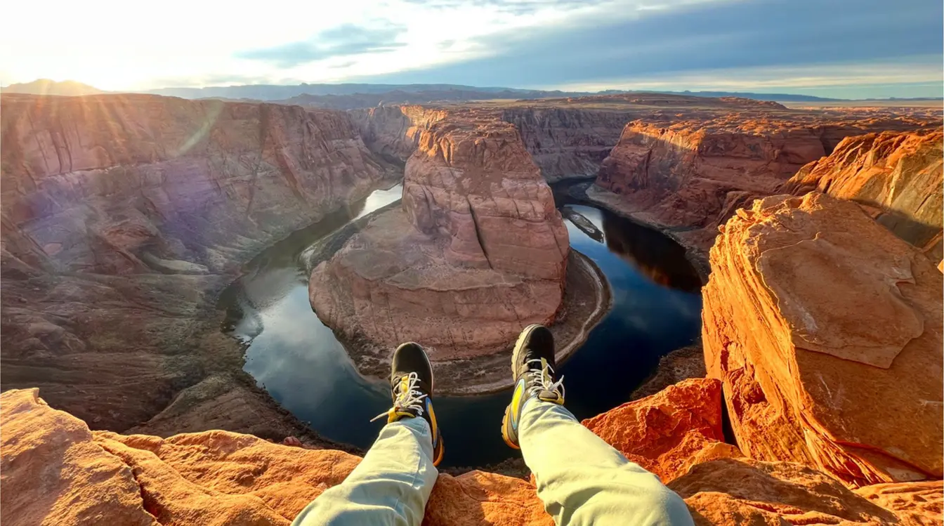 Una persona sentada en un acantilado mirando hacia un río en forma de herradura entre cañones de Grand Canyon, con luz de tarde.