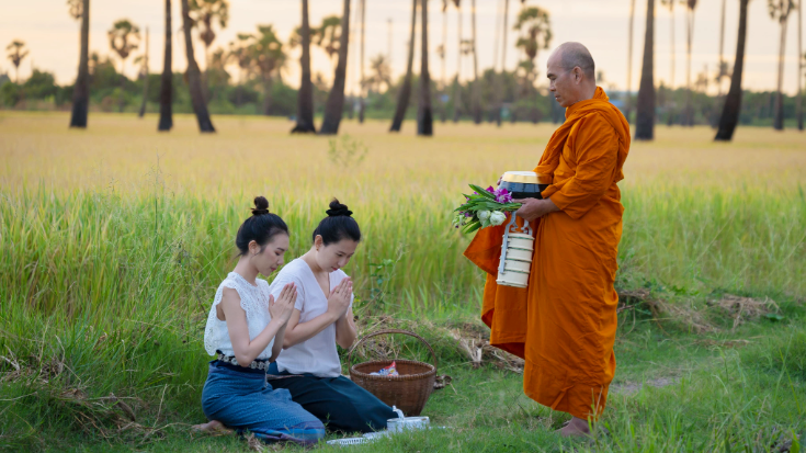 An image of a Thai monk walking the dirt road of a rural village in Thailand and practicing Pindapata, the tradition of accepting sustenance from local people to maintain the local communities of monks. Two women, dressed in white shirts and blue jeans, are kneeling before him. In the background is a green field with brown trees - to illustrate a blog post entitled '33 fun facts about Thailand'.