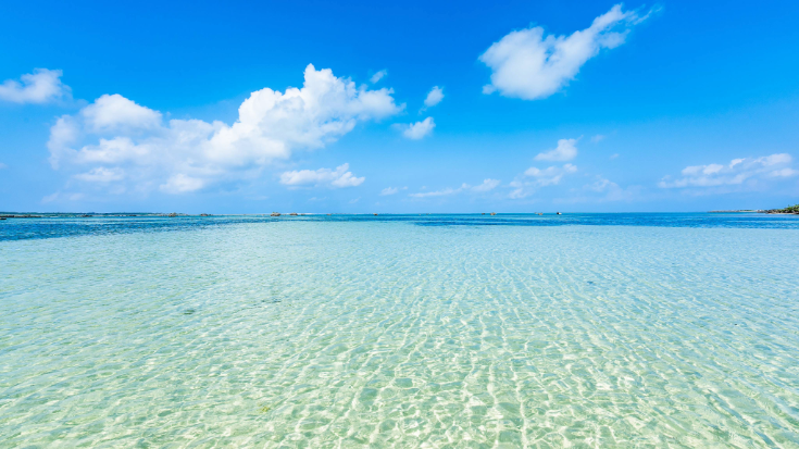 A colour photograph showing the shallow waters reflecting the bright sunshine on a white-sand beach on Miyako Island, Okinawa, Japan. In the foreground you can see the sand and the ocean water stretching out into the distance. overhead, the sky is bright blue with white clouds dotted through it. To illustrate a blog post entitled 'Top Things to Do in Okinawa, Japan's Tropical Gem.'