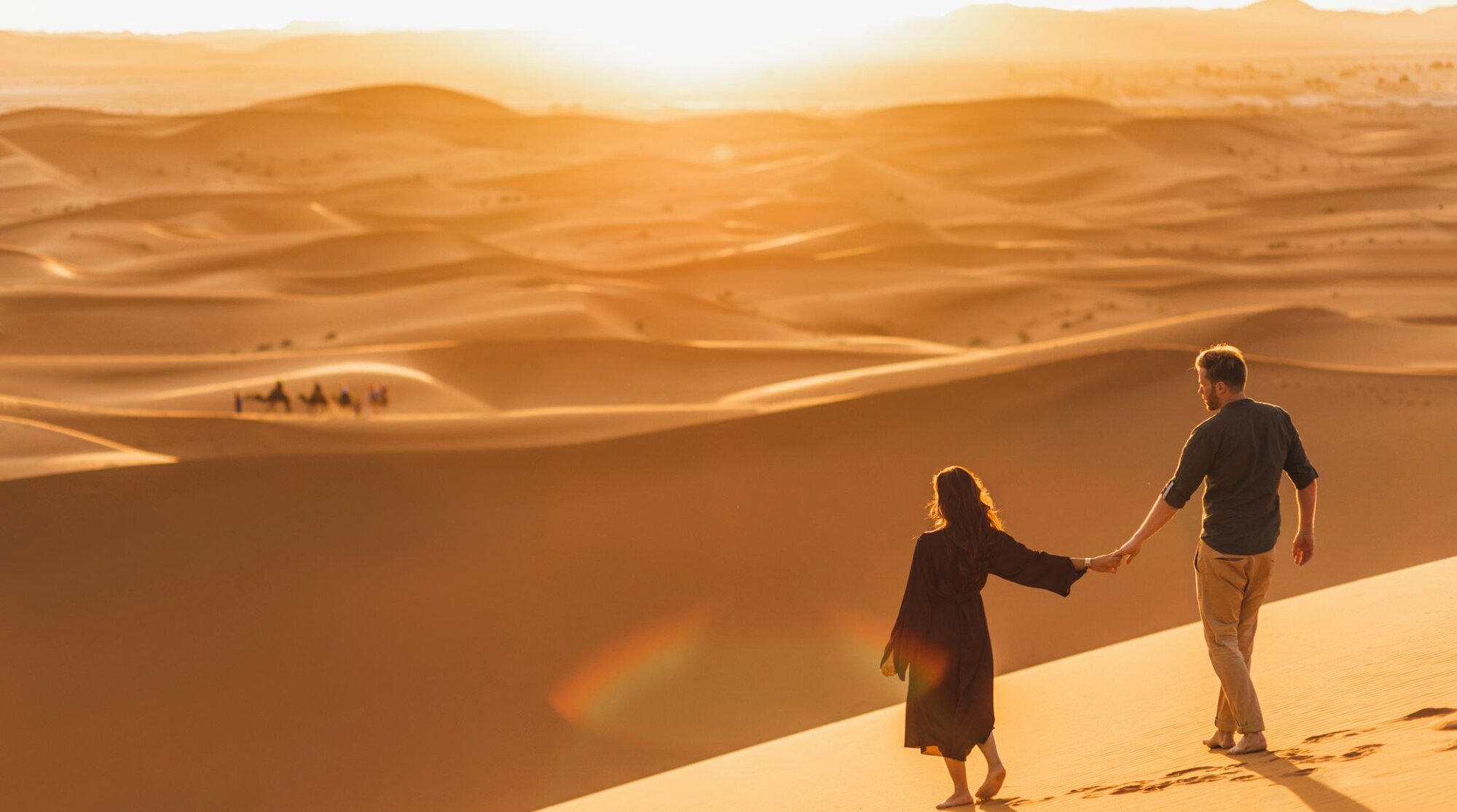Coppia che cammina nel deserto al tramonto, vista da dietro, durante un viaggio avventura con paesaggio sabbioso.