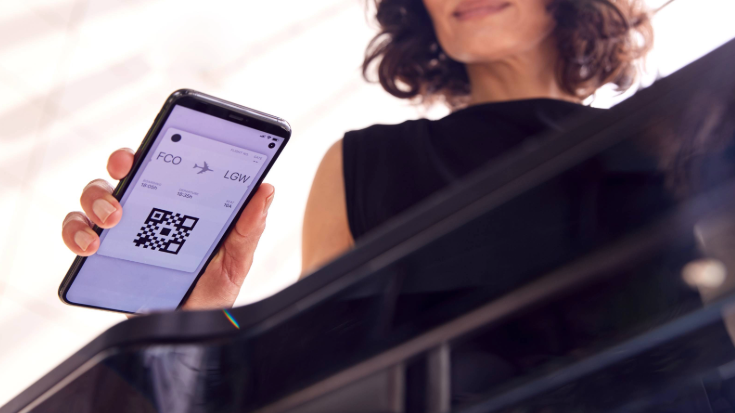 A photo of a woman with black hair in the background, in the foreground is a close-up of her mobile phone, which has a boarding pass on the screen for a flight from Rome Fiumicino Airport, heading to London Gatwick. She is holding the phone screen, which includes a QR code of her boarding pass, against a screen reader at the airport. To illustrate a blog post entitled 'What's the Difference Between the EES and ETIAS? A Guide for Brits.'