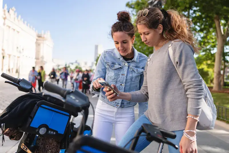 Dos amigas revisando un mapa en el celular junto a bicicletas en Madrid.