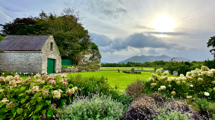 An Irish farmhouse sitting in the middle of a green field, surrounded by colourful flowers and a blue sky.