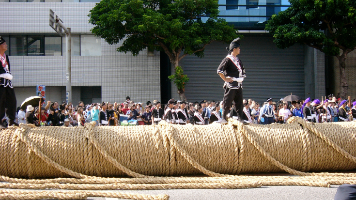 A colour photograph of the famous world record-breaking tug of war rope that is made by locals annually for the Naha Great Tug of War Festival. The rope is laid out along a standard residential Japanese street, with crowds lining it on either side. There is a man dressed in traditional Okinawan dress - black suit jacket and trousers with a white trim, standing on top of the rope looking away into the distance. To illustrate a blog post entitled 'Top Things to Do in Okinawa, Japan's Tropical Gem.'