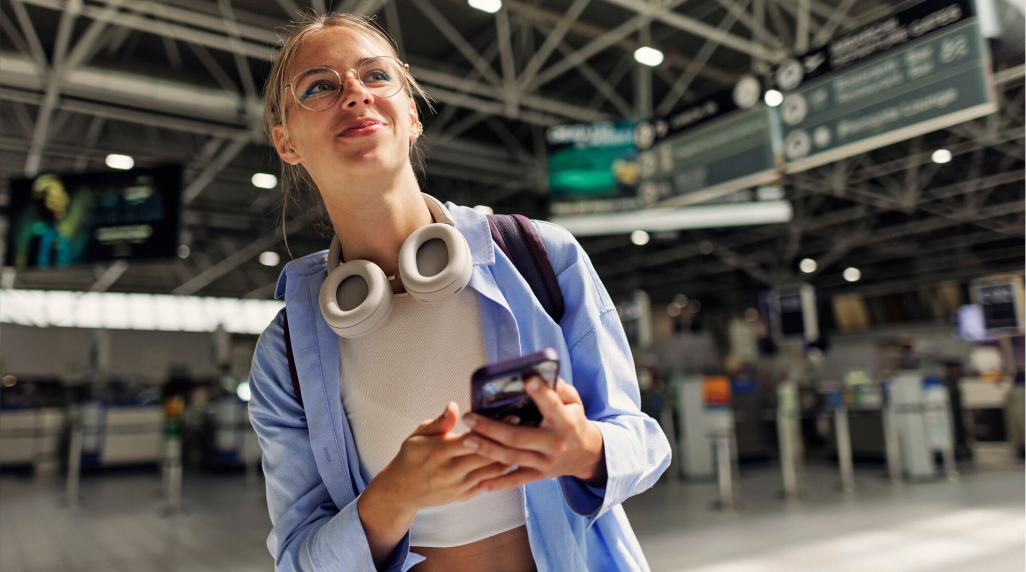 Jovem viajante com fone de ouvido no pescoço e mochila segurando celular no terminal do aeroporto.