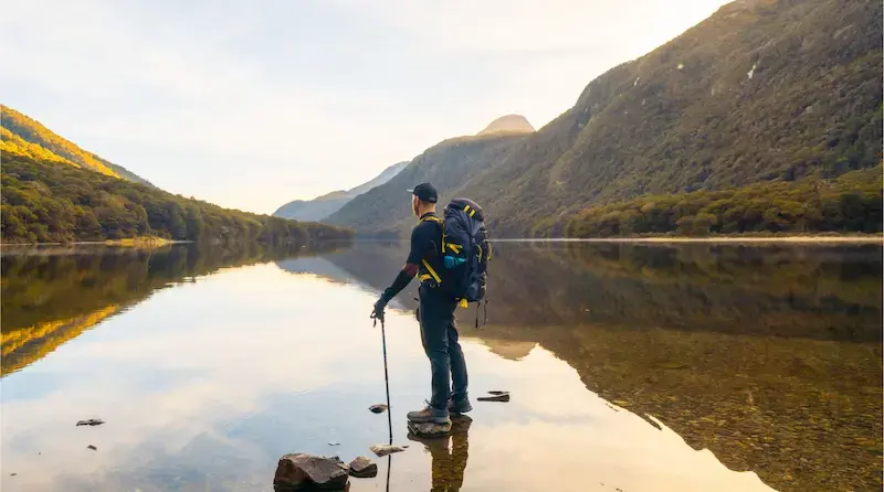 Mochilero de pie junto a un lago rodeado de montañas usando su teléfono durante un viaje