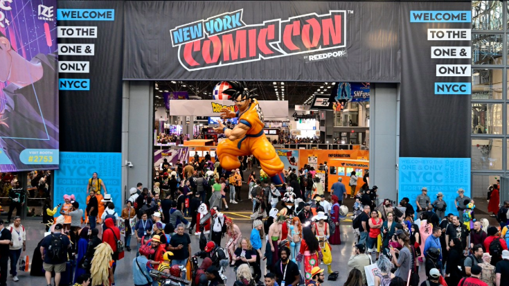 A photograph of a conference floor from above at the New York Comic Con event. There are lots of people walking around and an archway with a large black banner with letters in blue stating 'New York' and letters in red stating 'Comic Con'. In the middle of the arch, just underneath it, there is a large-scale manga character, a young man dressed in a yellow jumpsuit with blue arm bands and black hair. He is in a half-crouching pose, towering above the crowds.
