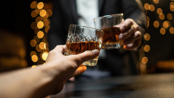 A colour image of two hands toasting with crystal patterned cut glasses filled about halfway with Scottish whiskey. To illustrate a blog post entitled 'What is Burns Night and How to Celebrate It'.