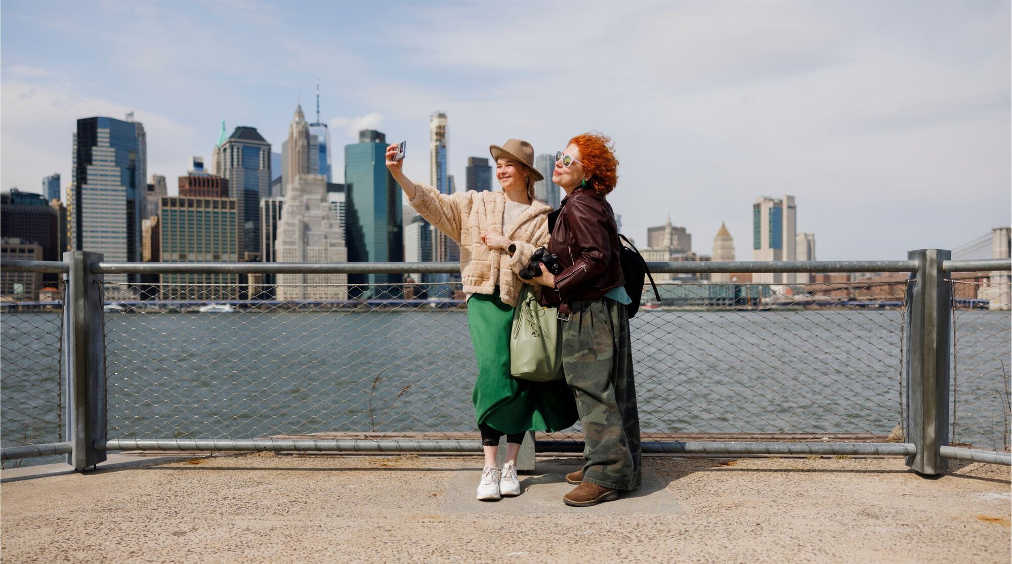 Turistas registrando uma selfie com o icônico horizonte de Manhattan e o Rio Hudson ao fundo.