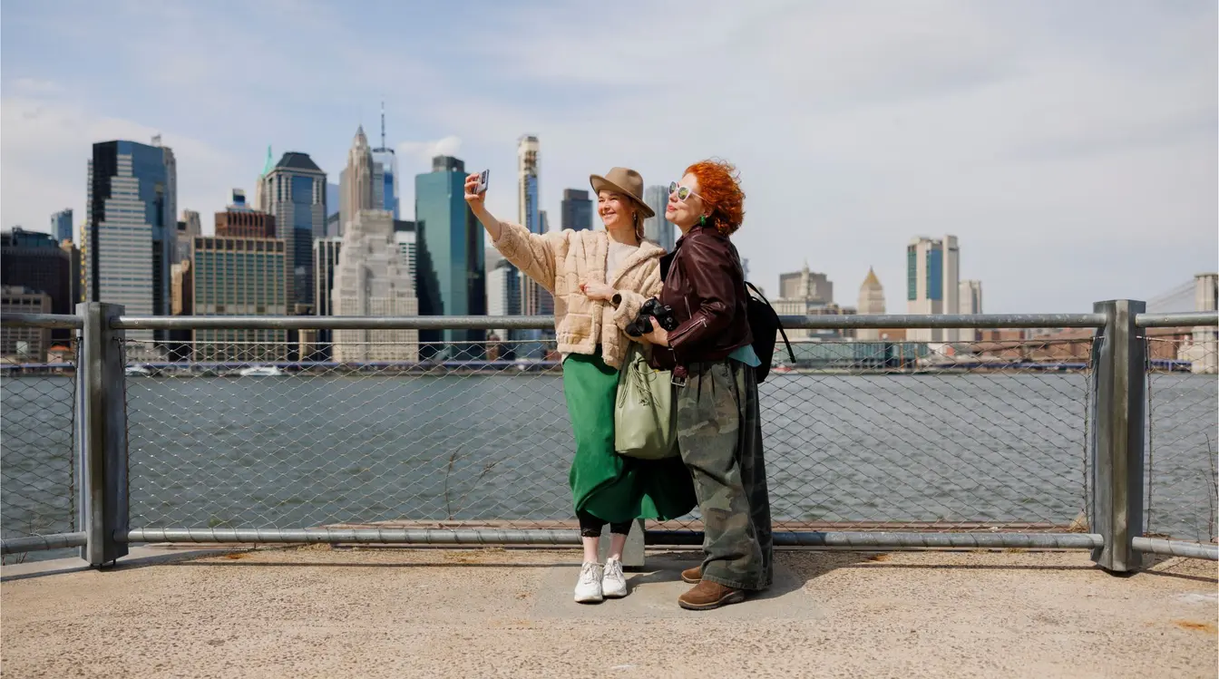 Turistas registrando uma selfie com o icônico horizonte de Manhattan e o Rio Hudson ao fundo.