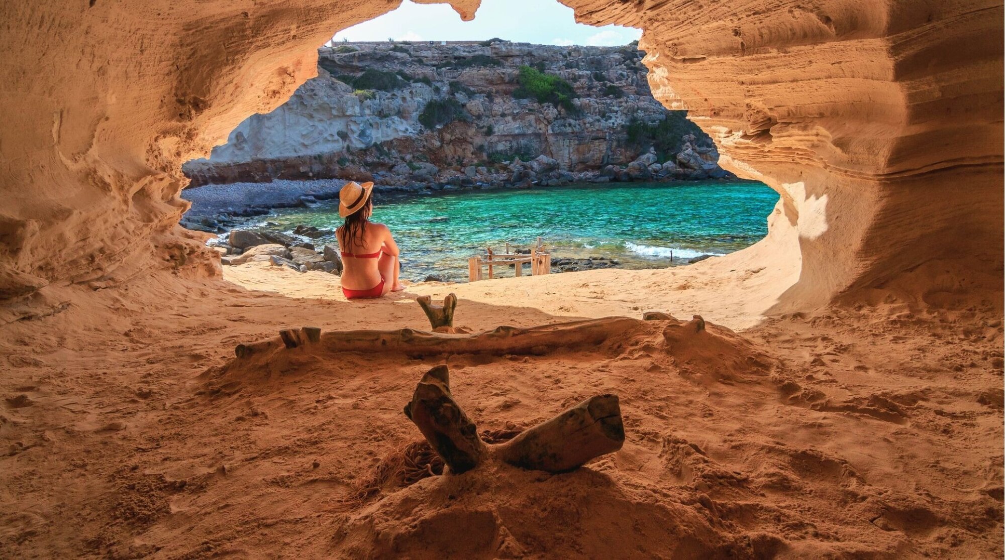 Una persona sentada mira una cala de agua turquesa desde el interior de una cueva junto al mar.