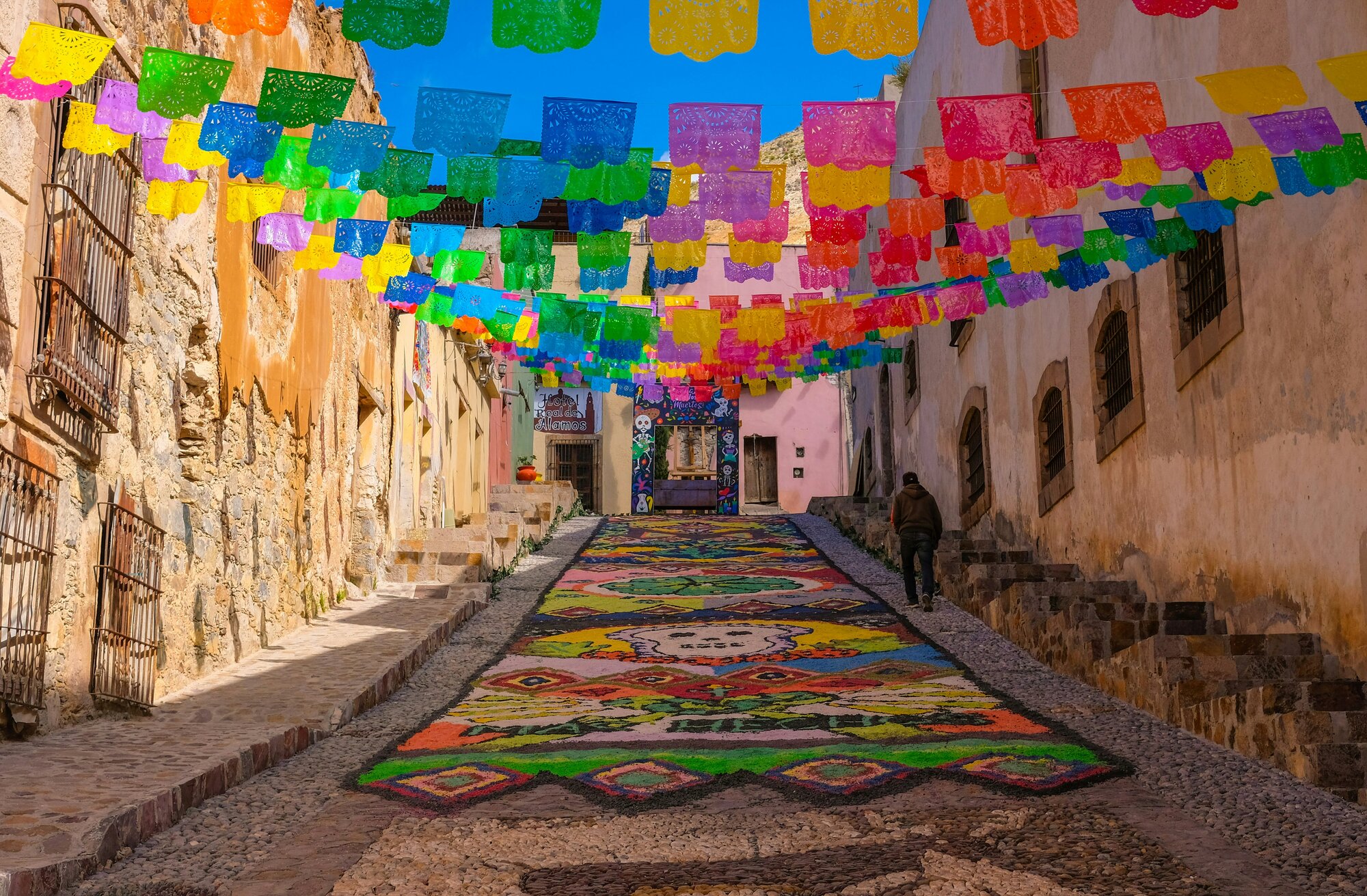 Imagen de una calle empedrada decorada con un tapete de arena y papel picado multicolor para ilustrar el Día de Muertos en Oaxaca