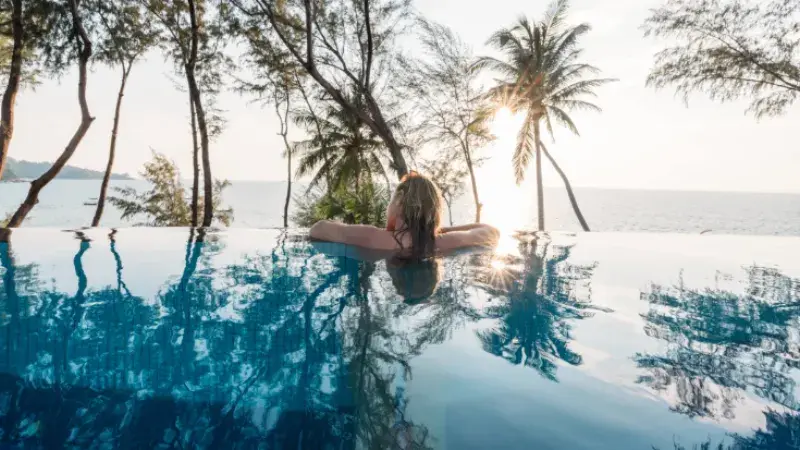 The back of a blonde-haired woman's head and arms as she looks out over the edge of an infinity pool at palm trees and sea. 