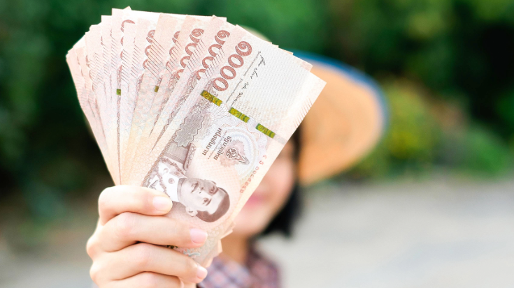 An image of some Thai Baht bank notes being held up to the camera close-up. A woman's hand is holding the notes and she is shown in the background blurred out - to illustrate a blog post entitled '33 fun facts about Thailand'.