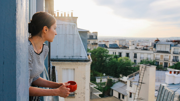 A woman wearing a black and white horizontally striped t-shirt holds a red mug while standing on a balcony and looking out over the rooftops of Montmartre, Paris.