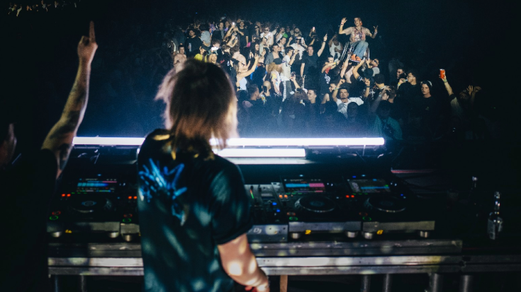 A photograph of a DJ in the foreground on is deck in the dark. In the background is a crowd dancing to and putting their hands in the air to his music. To illustrate a blog post entitled 'The Ultimate Sziget Festival Guide.'