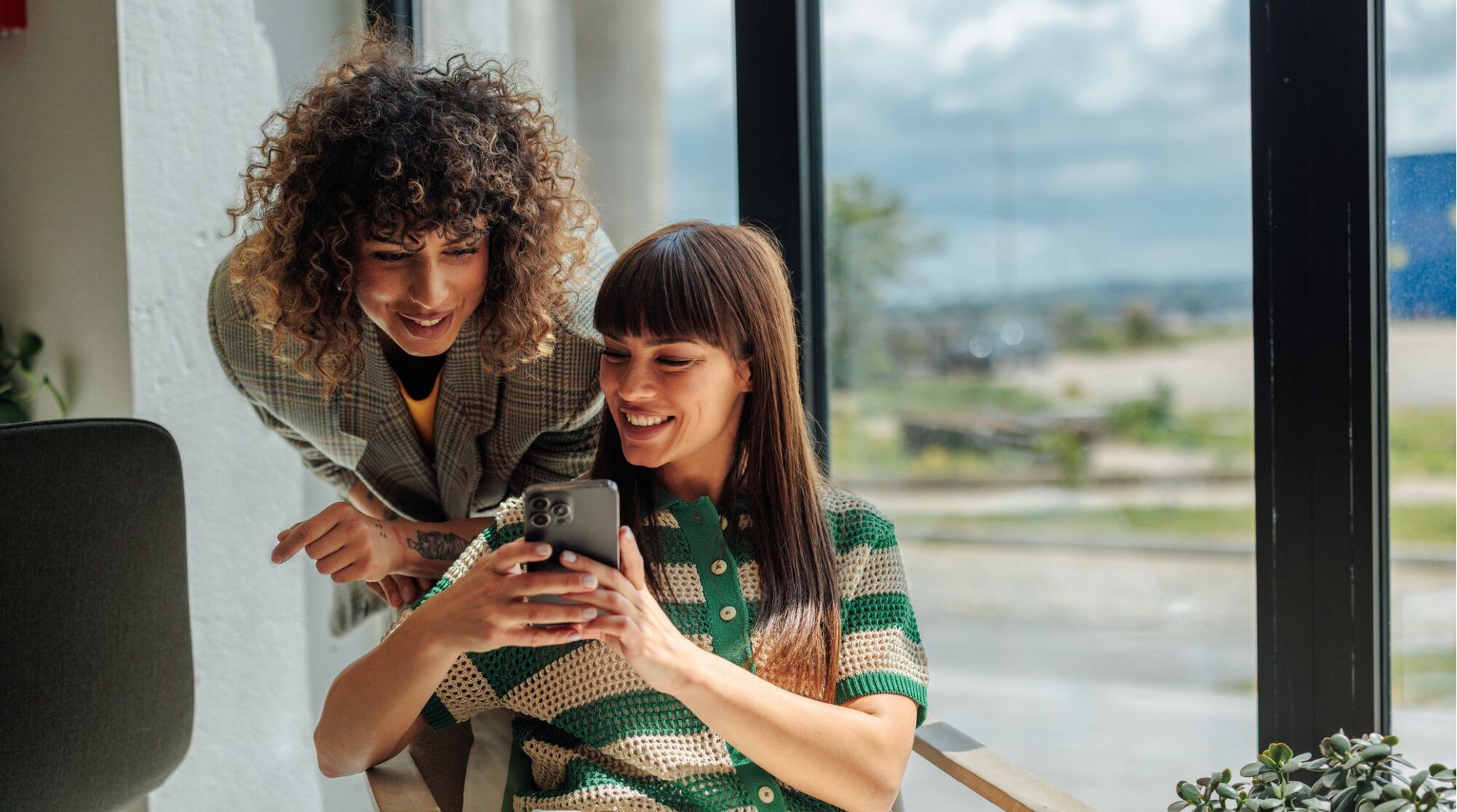 Duas colegas de trabalho alegres, compartilhando informações sobre chip físico ou eSIM, olhando para o celular no escritório.