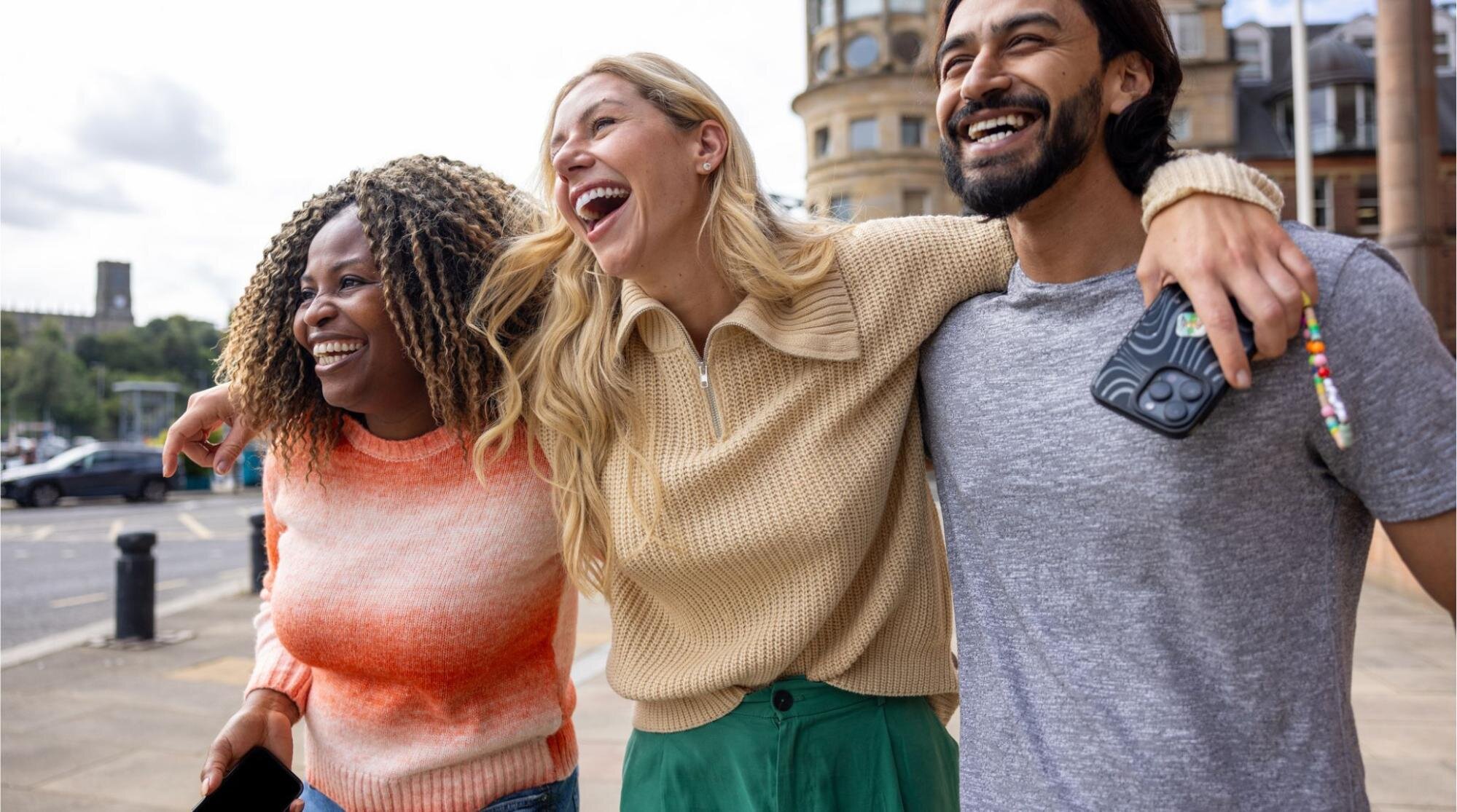Três pessoas caminhando abraçadas e sorrindo em uma rua da cidade. Uma delas segura um celular e se mantém conectada com chip internacional para Inglaterra.